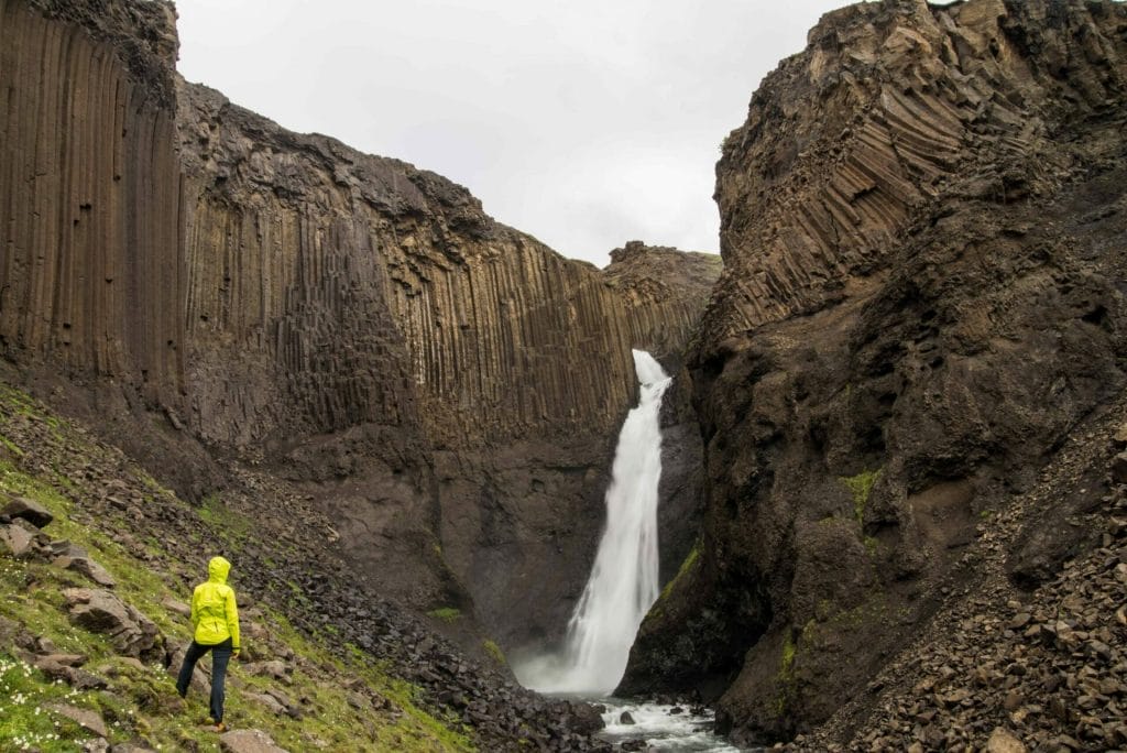 woman in yellow raincoat standing in front of Litlanesfoss basalt column waterfall in East Iceland