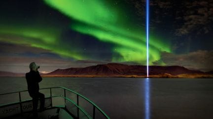 The Northern Lights shimmer in vibrant green hues above Reykjavik, framing the radiant glow of the Imagine Peace Tower on Viðey Island. The peaceful scene is captured from a yacht during a Northern Lights cruise, with the tower’s beam of light reaching upwards, blending with the auroras in the night sky. The calm waters reflect both the auroras and the Peace Tower’s illumination, creating a serene and magical atmosphere.
