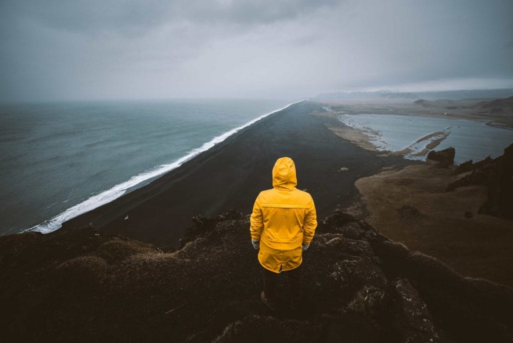 man in yellow raincoat watching over Reynisfjara black sand beach from Reynisfjall mountain