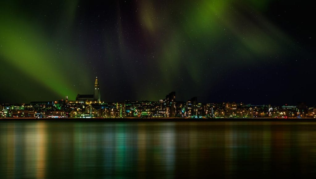 A stunning view of the Northern Lights illuminating the night sky above Reykjavik, as seen from a yacht during a Northern Lights cruise. The vibrant green auroras dance gracefully across the horizon, reflecting softly on the calm waters below, with the city’s lights twinkling in the background.