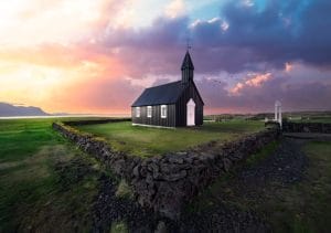 A stunning sunset view of Búðarkirkja, the iconic black church in Búðir, Snæfellsnes Peninsula, Iceland. The church stands boldly against a vibrant sky painted with hues of orange, pink, and purple, while the silhouette of the surrounding mountains adds depth to the scene. The church's dark exterior contrasts beautifully with the warm colors of the sunset, creating a serene and picturesque atmosphere that highlights the tranquil beauty of this historic site. Lush green fields surround the church, completing this enchanting landscape.