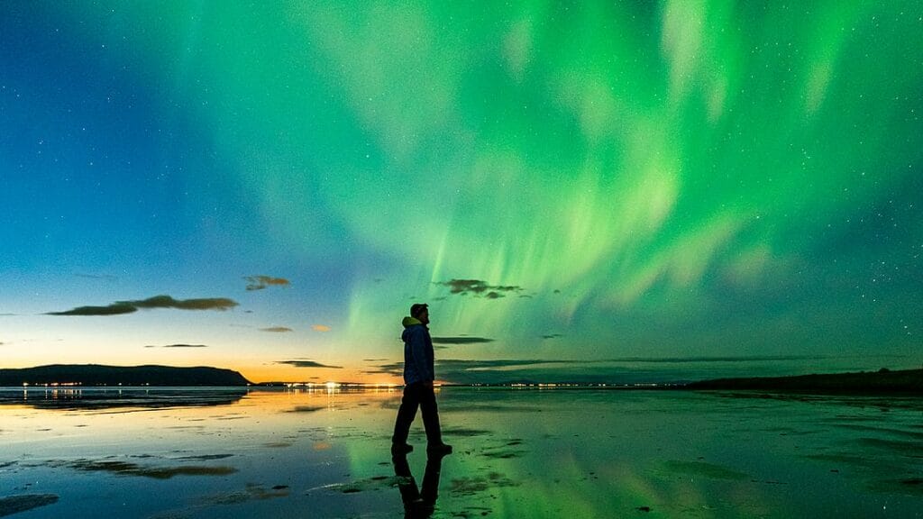 A captivating image from a Northern Lights photography tour, showcasing a group of photographers set against a stunning display of the Aurora Borealis. The sky is filled with vibrant greens and purples, swirling and shimmering above a snow-covered landscape. Each photographer is focused on capturing the ethereal beauty, with tripods set up and cameras aimed at the dazzling lights. In the background, the dark silhouettes of distant mountains add depth to the scene, creating an enchanting atmosphere as the night unfolds.