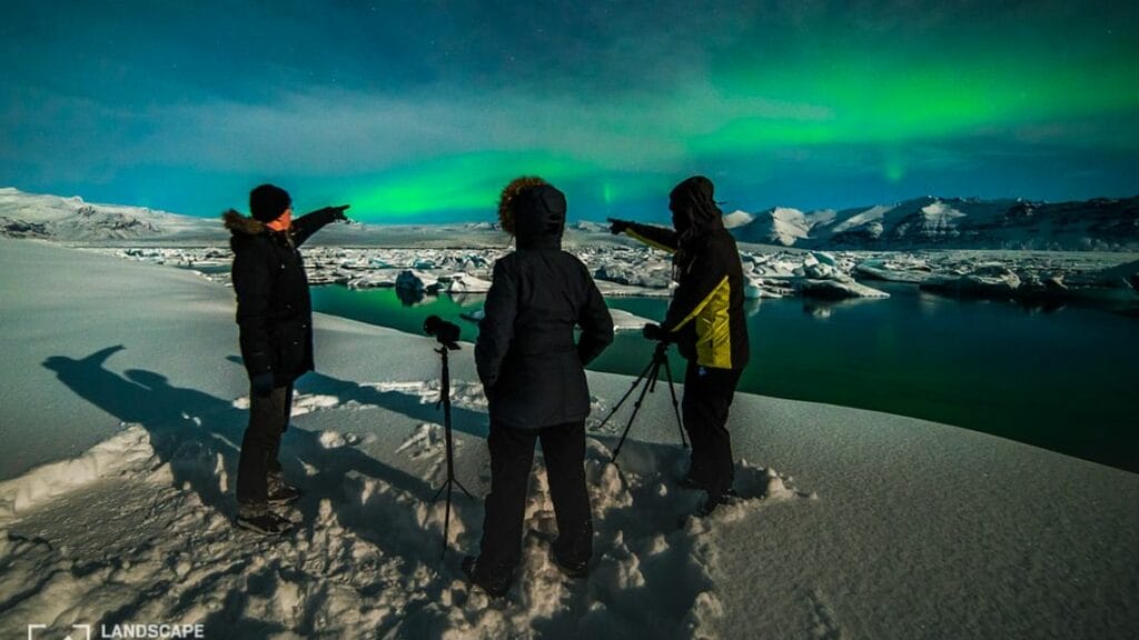A captivating image from a Northern Lights photography tour, showcasing a group of photographers set against a stunning display of the Aurora Borealis. The sky is filled with vibrant greens and purples, swirling and shimmering above a snow-covered landscape. Each photographer is focused on capturing the ethereal beauty, with tripods set up and cameras aimed at the dazzling lights. In the background, the dark silhouettes of distant mountains add depth to the scene, creating an enchanting atmosphere as the night unfolds.