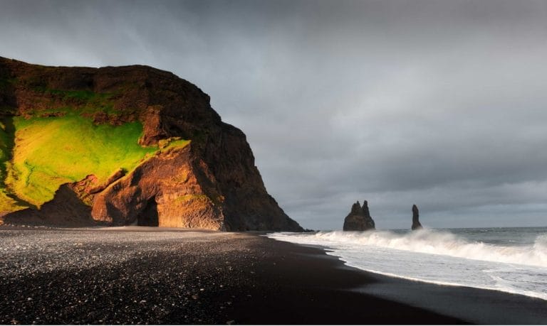 A breathtaking view of Reynisfjara black sand beach in Iceland, showcasing the striking Reynisdrangar cliffs in the background. The dark volcanic sand stretches across the foreground, while powerful waves crash against the shore. The towering, jagged cliffs rise majestically from the ocean, contrasting beautifully with the deep blue water and cloudy sky. The scene captures the wild and rugged beauty of Iceland's coastline.