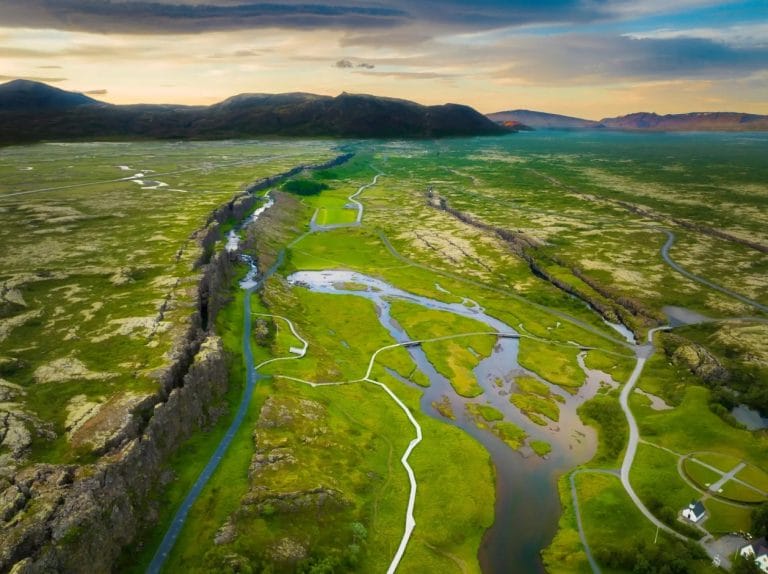 A scenic view of Þingvellir National Park in Iceland, showcasing the expansive rift valley where the North American and Eurasian tectonic plates meet. The photo captures the park's rugged landscape with jagged cliffs, patches of greenery, and a clear river winding through the valley. In the distance, volcanic mountains rise against a dramatic sky, while the Þingvallavatn lake shimmers under the soft light, adding to the park's natural beauty and historical significance.