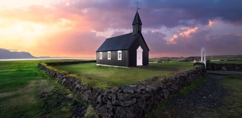 A stunning sunset view of Búðarkirkja, the iconic black church in Búðir, Snæfellsnes Peninsula, Iceland. The church stands boldly against a vibrant sky painted with hues of orange, pink, and purple, while the silhouette of the surrounding mountains adds depth to the scene. The church's dark exterior contrasts beautifully with the warm colors of the sunset, creating a serene and picturesque atmosphere that highlights the tranquil beauty of this historic site. Lush green fields surround the church, completing this enchanting landscape.