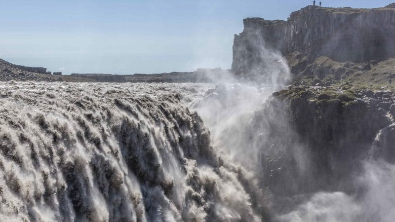 A stunning view of Dettifoss waterfall in Iceland, showcasing its powerful cascade as it plunges dramatically into a rugged canyon. The waterfall, known as Europe's most powerful, creates a misty spray that catches the sunlight, forming a rainbow in the air. Surrounding the waterfall, steep cliffs and rocky terrain highlight the natural beauty and raw power of this iconic landmark, while the lush greenery in the distance adds a touch of color to the scene.