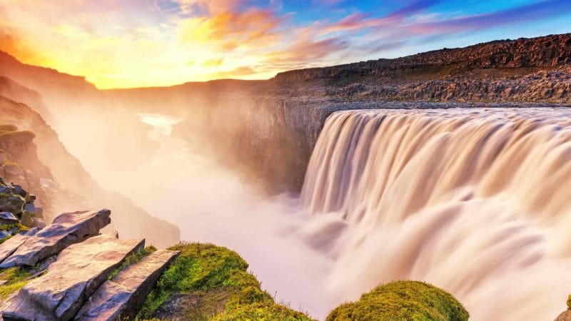 A breathtaking sunset casts warm hues of orange, pink, and purple over Dettifoss waterfall in Iceland. The cascading water glows softly in the fading light as it plunges dramatically into the rocky gorge below. Wisps of mist rise from the falls, catching the last rays of sunlight, while the surrounding landscape, dotted with rugged rocks and greenery, creates a serene backdrop. The sky transitions into twilight, highlighting the natural beauty and tranquility of this iconic location.