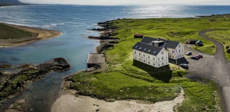 An aerial view of Hotel Búðir, beautifully situated on the Snæfellsnes Peninsula in Iceland. The hotel is surrounded by lush green landscapes and rugged mountains, with the iconic black Búðakirkja church visible nearby. The expansive view highlights the stunning contrast between the dark, volcanic terrain and the vibrant greenery, while the majestic Snæfellsjökull glacier looms in the background. The image captures the serene and tranquil setting of the hotel, showcasing its ideal location for enjoying the breathtaking natural beauty of the region.
